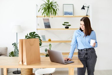 Kız masada duruyor, dizüstü bilgisayara bakıyor, bir bardak içki tutuyor, ve masanın yanında kağıt alışveriş torbaları var.