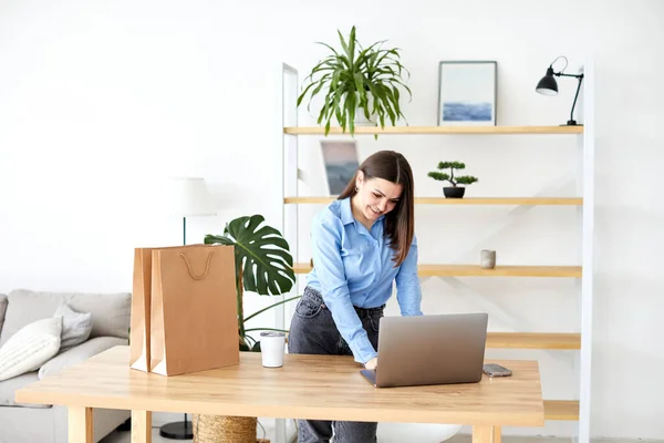 Happy businesswoman working in a home office standing looking at her laptop computer surrounded by gift bags and packaging