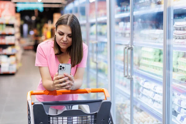 Happy caucasian woman using mobile phone in grocery shopping. Checklist application buying food products in supermarket. A housewife in a grocery store makes purchases while standing next to a cart and writes a message on the phone