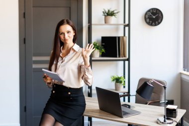 Happy business woman using pen and writing something in her notebook, raising her hand up, showing a greeting gesture