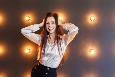 Pretty brunette in blouse and short skirt stands in studio with brick wall and lamps and keeps his hands on his head