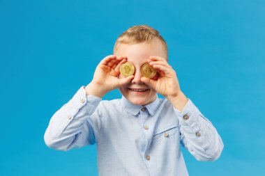 Kripto para ve modern finans. Neşeli çocuk stüdyoda mavi arka planda altın Bitcoin tutuyor ve gözlerini bozuk paralarla kapatıyor.