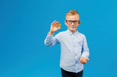 Kripto para ve modern finans. Gözlüklü neşeli çocuk mavi arka planda stüdyoda altın bitcoin tutuyor. Geleceğin iş adamı.