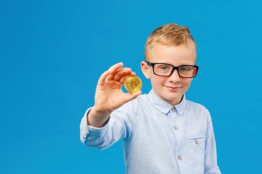 Kripto para ve modern finans. Gözlüklü neşeli çocuk mavi arka planda stüdyoda altın bitcoin tutuyor. Geleceğin iş adamı.