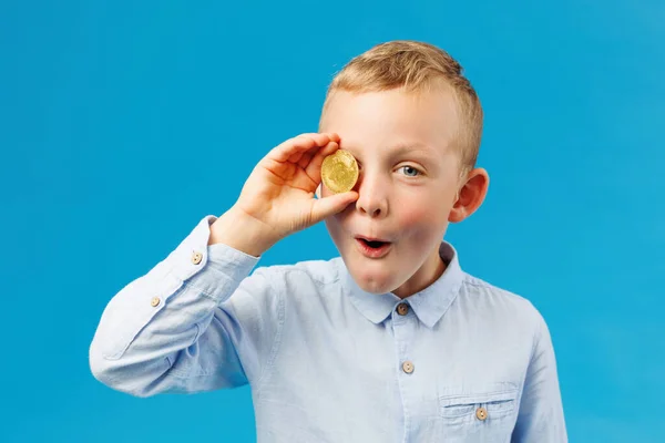Cryptocurrency and modern finance. Cheerful boy holding golden bitcoin in studio on the blue background and covers his eye with a coin