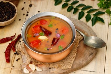 Rasam, Güney Hintlilerin ana öğesi, baharatlı-tatlı-ekşi bir sebze çorbası geleneksel olarak pirinç bir kaba konur, yanında malzemeler, ahşap desenli arka plan.
