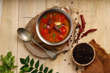 Rasam, Güney Hintlilerin ana öğesi, baharatlı-tatlı-ekşi bir sebze çorbası geleneksel olarak pirinç bir kaba konur, yanında malzemeler, ahşap desenli arka plan.