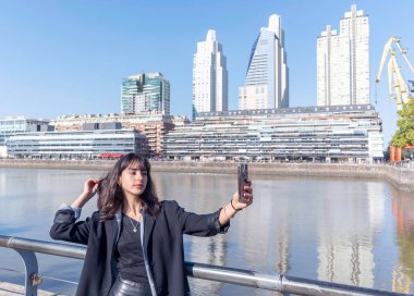 Güneşli bir günde limanda akıllı telefon kullanan genç bir kadın selfie çekiyor. Boşluğu kopyala