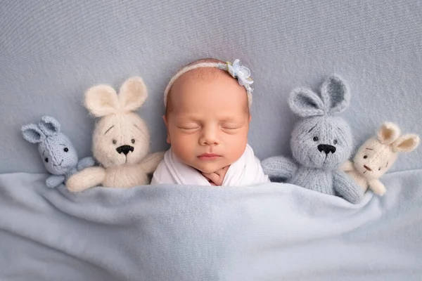 Спящая новорожденная девочка в первые дни жизни в белом коконе с белой повязкой и голубым цветком на синем фоне. Студийная макрофотография, портрет новорожденной балерины, игрушки-кролики. — стоковое фото