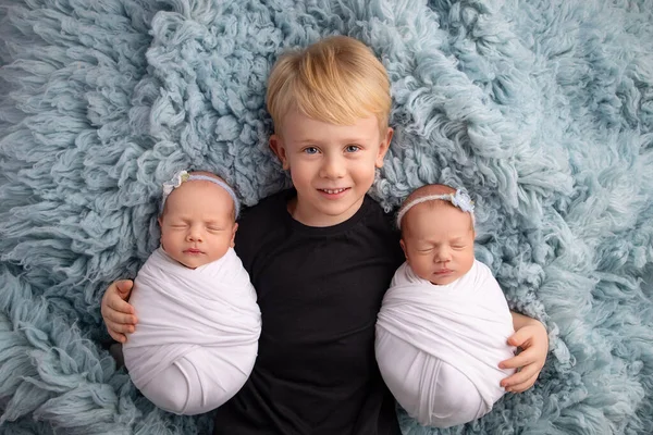 Drobná novorozená dvojčata v bílých zámotcích na modrém pozadí. Starší bratr leží mezi nimi a něžně objímá své dvojče. Novorozená dvojčata v bílých čelenkách s bílými a modrými květy — Stock fotografie