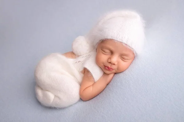 Dormir niña recién nacida en los primeros días de vida en un traje de cuerpo blando blanco con una gorra blanca de lana de punto sobre un fondo azul. Estudio macrofotografía, retrato de un recién nacido. Mujeres felicidad. — Foto de Stock