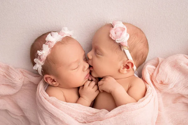Pequeñas gemelas recién nacidas. Un gemelo recién nacido duerme junto a su hermana. Niñas gemelas recién nacidas en el fondo de una manta rosa con vendajes rosas. Las chicas suavemente abrazan y besan a su hermana en una pose linda. — Foto de Stock