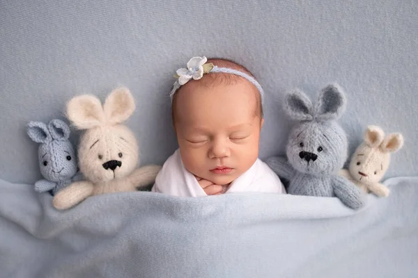 Niña recién nacida dormida en los primeros días de vida en un capullo blanco con una venda blanca y una flor azul sobre un fondo azul. Estudio macro fotografía, retrato de una bailarina recién nacida, juguetes conejitos. — Foto de Stock