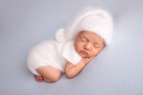 Slapen pasgeboren meisje in de eerste dagen van het leven in een witte zachte bodysuit met een gebreide wollen witte pet op een blauwe achtergrond. Studio macro fotografie, portret van een pasgeborene. Vrouwen geluk. — Stockfoto