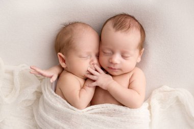 Tiny newborn twins boys in white cocoons on a white background. A newborn twin sleeps next to his brother. Newborn two twins boys hugging each other.Professional studio photography