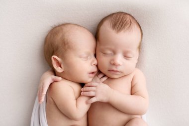 Tiny newborn twins boys in white cocoons on a white background. A newborn twin sleeps next to his brother. Newborn two twins boys hugging each other.Professional studio photography