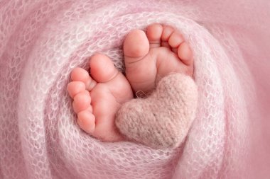 Knitted pink heart in the legs of a baby. Soft feet of a new born in a pink wool blanket. Close-up of toes, heels and feet of a newborn. Macro photography the tiny foot of a newborn baby.