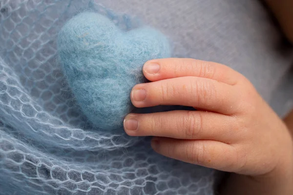 Tiny hand of a newborn. The soft hands of a newborn hold a blue woolen heart. The fingers of the hands of a newborn close-up. Studio macro photo on the background of a blue blanket.