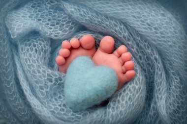 Tiny foot of a newborn. Soft feet of a newborn in a turquoise woolen blanket. Close-up the toes, heels and feet of a newborn baby. Knitted turquoise heart in the legs of a baby. Macro photography. 