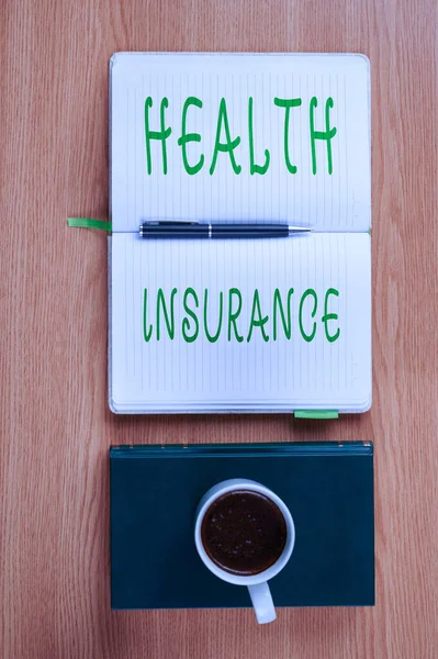 テキスト健康保険を表示する書き込み。医療費や手術費を支払うカバーを意味する概念プレーンブランクノートブックとコーヒーのカップの横にペン配置ブックテーブル. — ストック写真