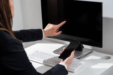 İş kadını cep telefonunu tutuyor ve bilgisayar ekranını işaret ediyor..