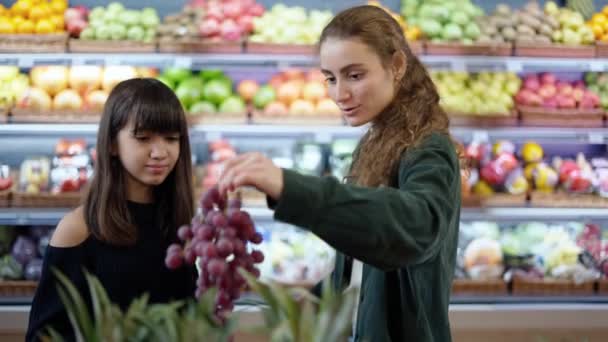 Dos muchachas alegres escogiendo las frutas biológicas - la uva en el supermercado juntos — Vídeos de Stock