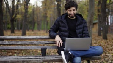Kamu parkında sıradan bir şekilde oturan gülümseyen adam video araması için dizüstü bilgisayar kullanıyor.
