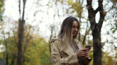 Gülümseyen kadın mesajları okuyor ya da sonbahar parkında telefondan internette sörf yapıyor.