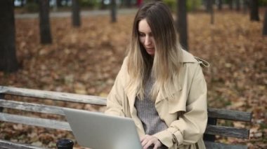 Genç ve güzel bir kadın dizüstü bilgisayarda çalışıyor, elinde kahveyle bankta oturuyor.