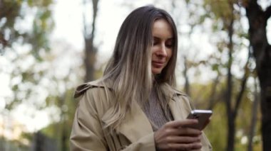 Gülümseyen kadın mesajları okuyor ya da sonbahar parkında telefondan internette sörf yapıyor.