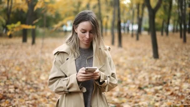 Junge blonde Frau schreibt Nachricht auf Handy in einem Herbstpark — Stockvideo