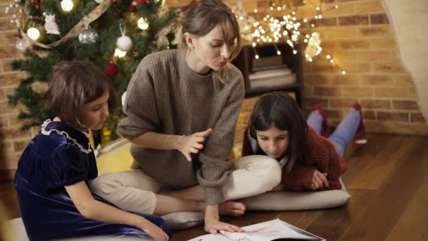 Мама с детьми сидит на полу читать книгу вместе на заднем плане Рождественские гирлянды огни — стоковое видео