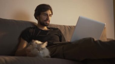 Adam evde dizüstü bilgisayarda çalışırken kanepede oturup bir kediyi okşuyor.