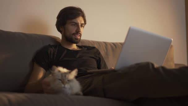 Ο άνθρωπος που εργάζεται σε ένα φορητό υπολογιστή στο σπίτι, ενώ κάθεται στον καναπέ και χαϊδεύει μια γάτα — Αρχείο Βίντεο