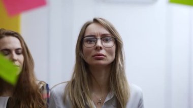 Takım toplantısındaki sarışın iş kadını cam duvara renk notu yapıştırıyor.