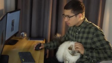 Genç adam ev ofisinden ekranlara bakıyor. Beyaz tüylü sevimli bir kediyle oturuyor.
