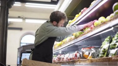 Pazarlamacı marketin meyve reyonunda raflara elma koymakla meşgul, sakallı adam önlük giyiyor.