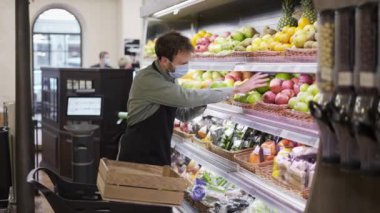 Maskeli bir satıcı marketin meyve reyonundaki raflara elma koymakla meşgul. Sakallı adam önlük takıyor.