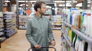 Adam süpermarkette yürüyor ve evdeki kimyasallar bölümünün raflarından bir şeyler alıyor.