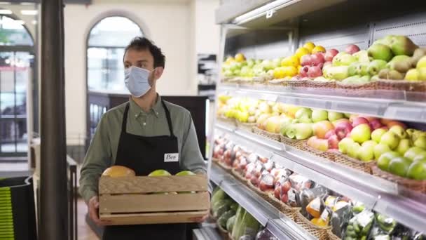 Portrait d'un bel homme en masque facial et tablier allant de l'avant dans un magasin d'aliments avec boîte de fruits à l'intérieur. — Video