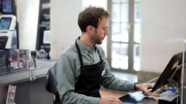 Markette çalışan tezgahtar. Kasada siyah önlüklü, gülümseyen erkek kasiyer ve tanınmayan bir müşteri.