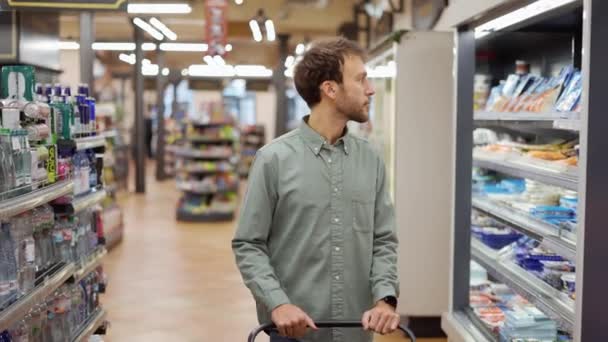 Homem feliz na camisa andando com um carrinho através do supermercado escolhendo mantimentos — Vídeo de Stock