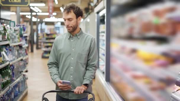 Homem feliz de camisa andando com um carrinho através do supermercado escolhendo mantimentos. Batendo a lista de compras no celular. — Vídeo de Stock