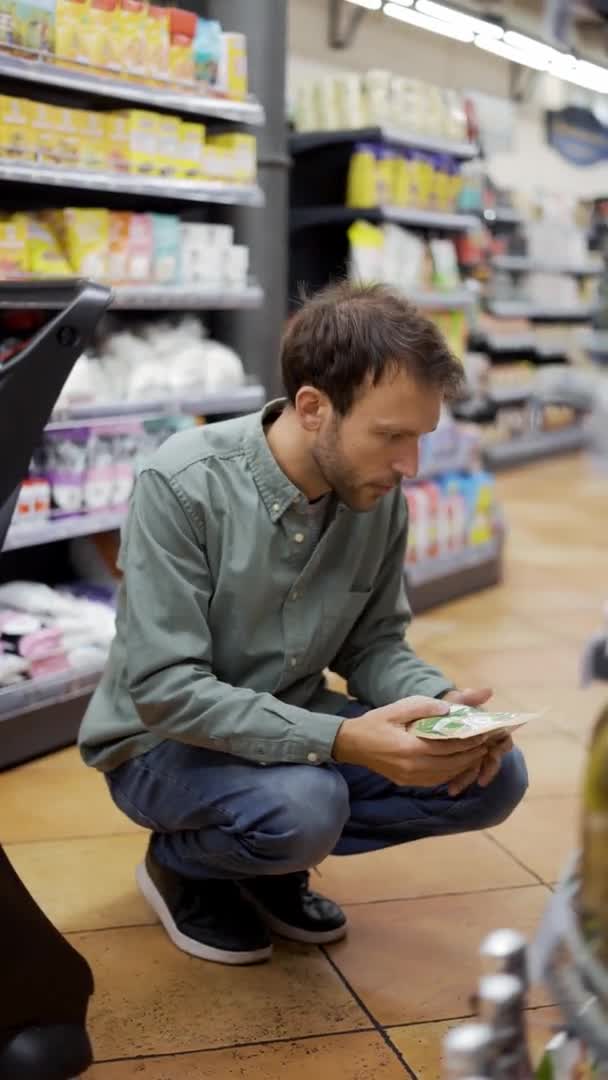 Boldog férfi ingben ül egy bevásárlókocsi mellett, és élelmiszert választ. Függőleges felvétel. Hogy egy guggolás, hogy vegye le az alsó polcon