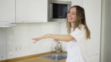 Mutfakta beyaz pijamalarıyla dans eden ve kulaklıkla müzik dinleyen mutlu genç bir kadın.
