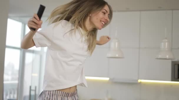 Divertida rubia juguetona bailando y cantando en casa divirtiéndose, use el mando a distancia como micrófono — Vídeos de Stock