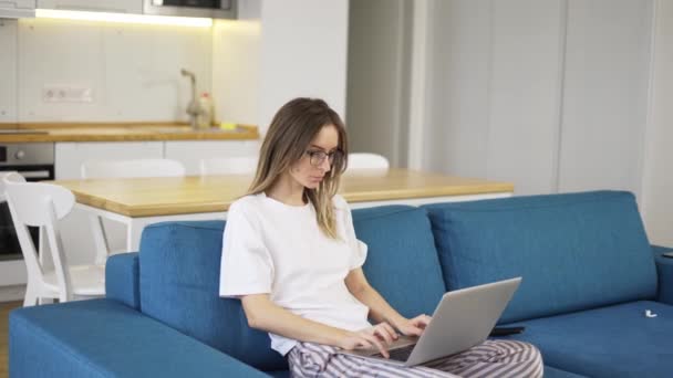 Blonda femeie în pijama sta pe canapea și tastând pe laptop de argint — Videoclip de stoc