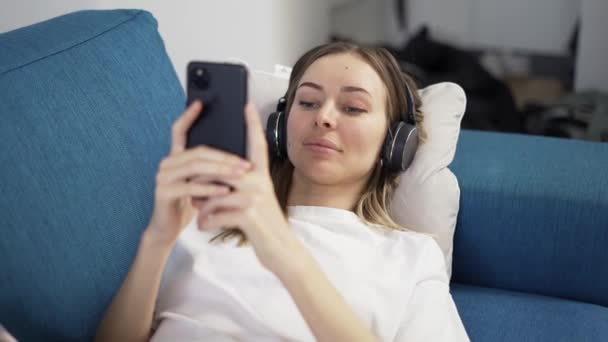 リラックスしたブロンドの女性は、携帯電話を使用してヘッドフォンで音楽を聴くソファ枕に横たわっている — ストック動画