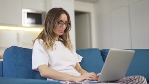 Femme blonde en pyjama assise sur le canapé et tapant sur un ordinateur portable en argent, vue basse — Video