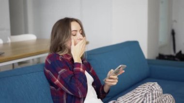 Evde koltukta oturmuş akıllı telefonları kontrol eden sıkıcı bir kadın.